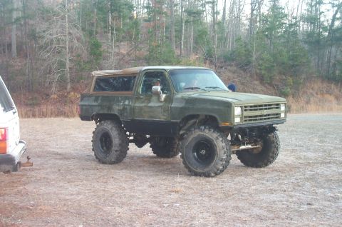 1987 Ford Bronco - Modified green SUV with oversized tires sits off-road, with another vehicle partially visible on the left.