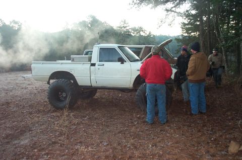 1987 Ford Bronco - Several people stand around a modified white pickup truck emitting smoke on a wooded dirt road.