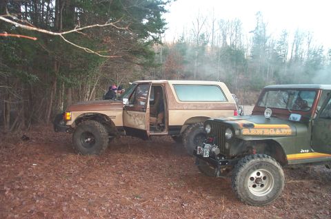 1987 Ford Bronco - A brown and tan 1987 Ford Bronco is parked off-road next to a Jeep, with trees and fallen leaves in the background.