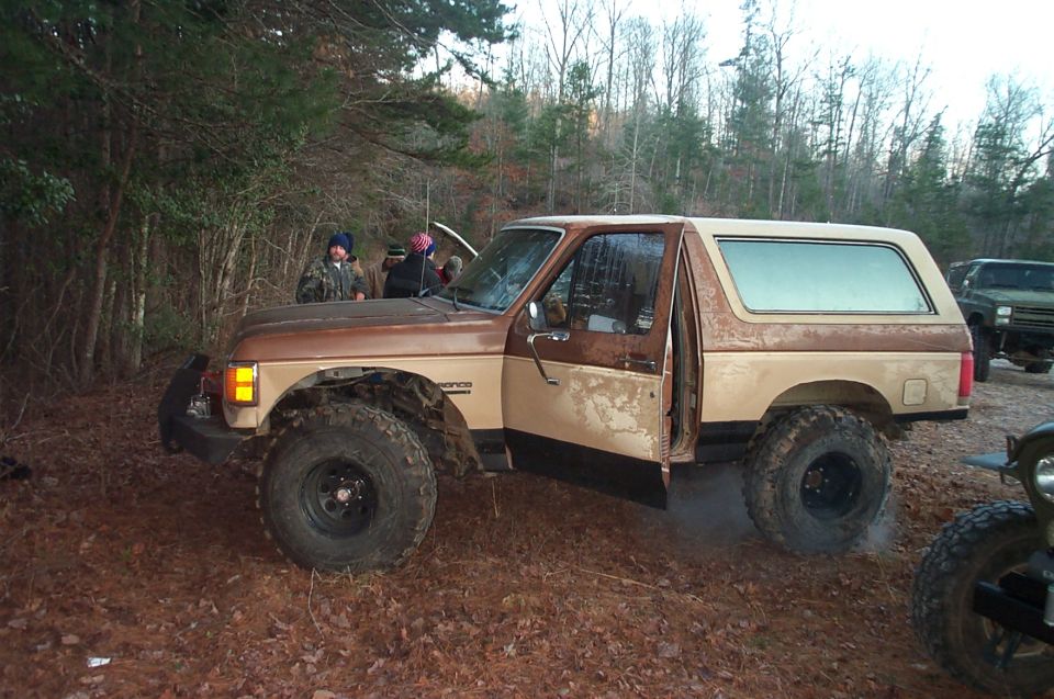 1987 Ford Bronco with lifted suspension and two-tone paint, parked in a wooded area with people around