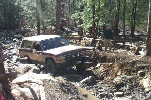 1987 Ford Bronco - A 1987 Ford Bronco is seen off-roading through a challenging, rocky, and muddy trail with a person observing nearby.