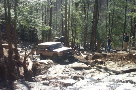 1987 Ford Bronco - 1987 Ford Bronco driving through a challenging rocky stream in a wooded area. Another vehicle is parked behind it.