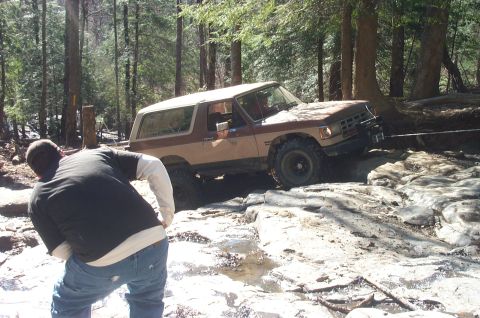 1987 Ford Bronco - A 1987 Ford Bronco is being winched through a challenging off-road course in a wooded area.