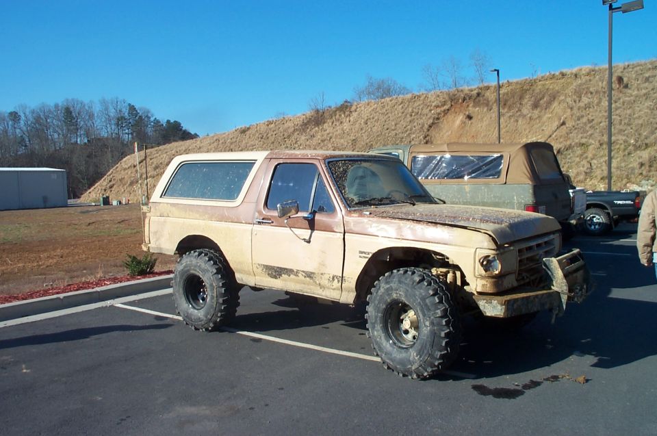 Tan and brown Ford Bronco with large off-road tires, dented front bumper, and visible rust