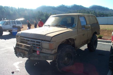 1987 Ford Bronco - Muddy 1987 Ford Bronco parked outdoors with other vehicles and people in the background.