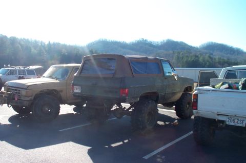 1987 Ford Bronco - Multiple off-road vehicles are parked in a lot, with a person visible working on one of them.