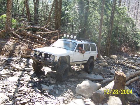 1987 Ford Bronco - A white Jeep Cherokee XJ is shown off-roading on a rocky trail, equipped with auxiliary lights on its roof rack.