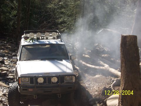1987 Ford Bronco - Jeep Cherokee XJ navigates a difficult off-road path with fallen logs and smoke.