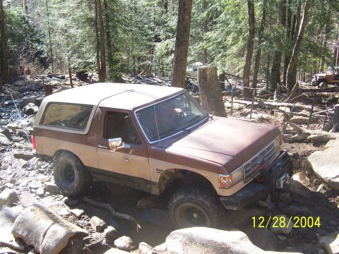 1987 Ford Bronco - A two-tone Ford Bronco navigates a challenging, rocky terrain in a wooded area.