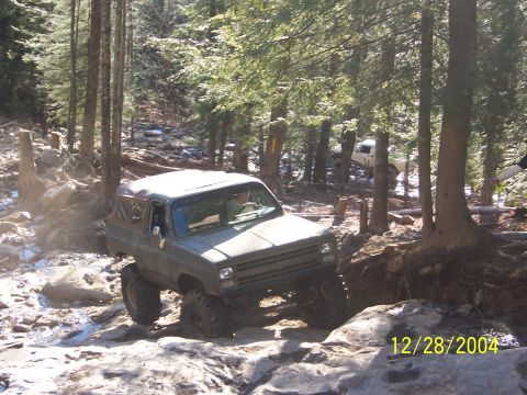 1987 Ford Bronco - An off-road SUV tackles a challenging rocky terrain in a wooded area.