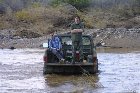 1994 Ford Bronco - Two men stand and sit in the bed of a pickup truck as it drives through a shallow river.
