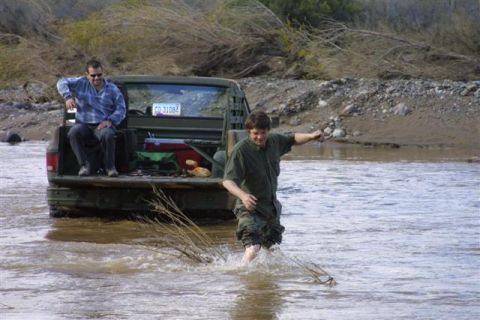 1994 Ford Bronco - Two men are in and around a green pickup truck partially submerged in a river, with one man sitting in the truck bed and the other wading t…