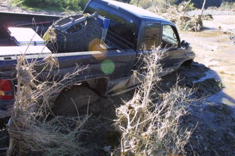 1994 Ford Bronco - A black pickup truck is stuck in a muddy riverbed, with vegetation obscuring parts of the vehicle.