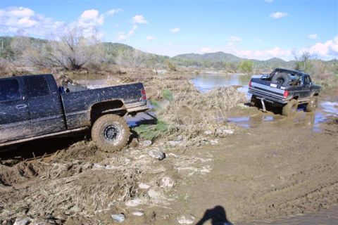 1994 Ford Bronco - Two trucks are stuck in a muddy, watery area with hills and trees in the background.