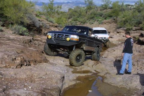 1994 Ford Bronco - Two off-road vehicles navigate a rocky, wet terrain with a man observing the action.