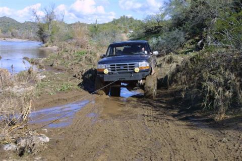 1994 Ford Bronco - A dark blue Ford Bronco navigates a muddy trail near a river, its front tires deep in the mire.