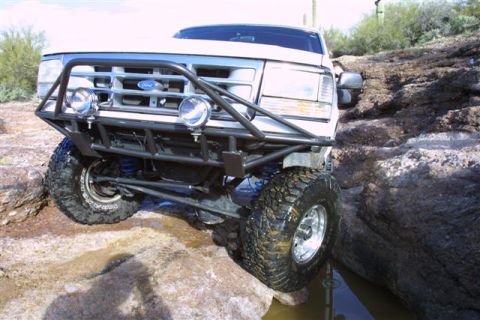 1994 Ford Bronco - A white Ford Bronco is shown off-roading on a rocky path, its front end angled towards the viewer.