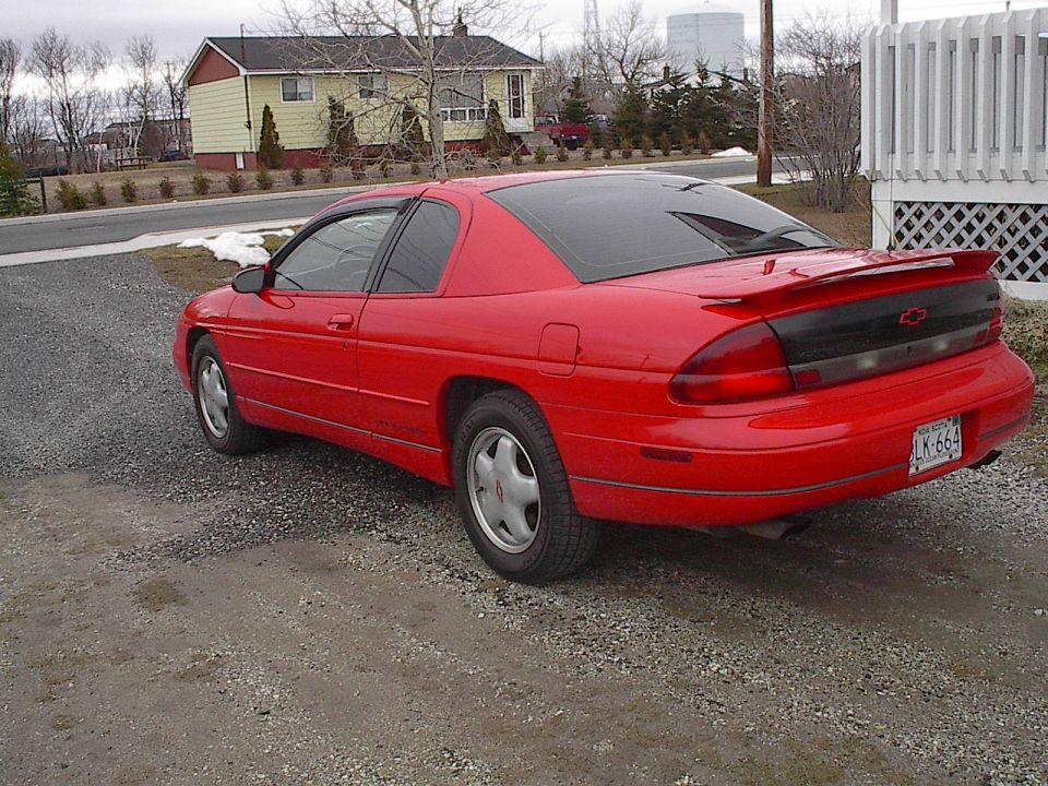 Red Chevrolet Monte Carlo coupe rear three-quarter parked on gravel by a white fence and houses