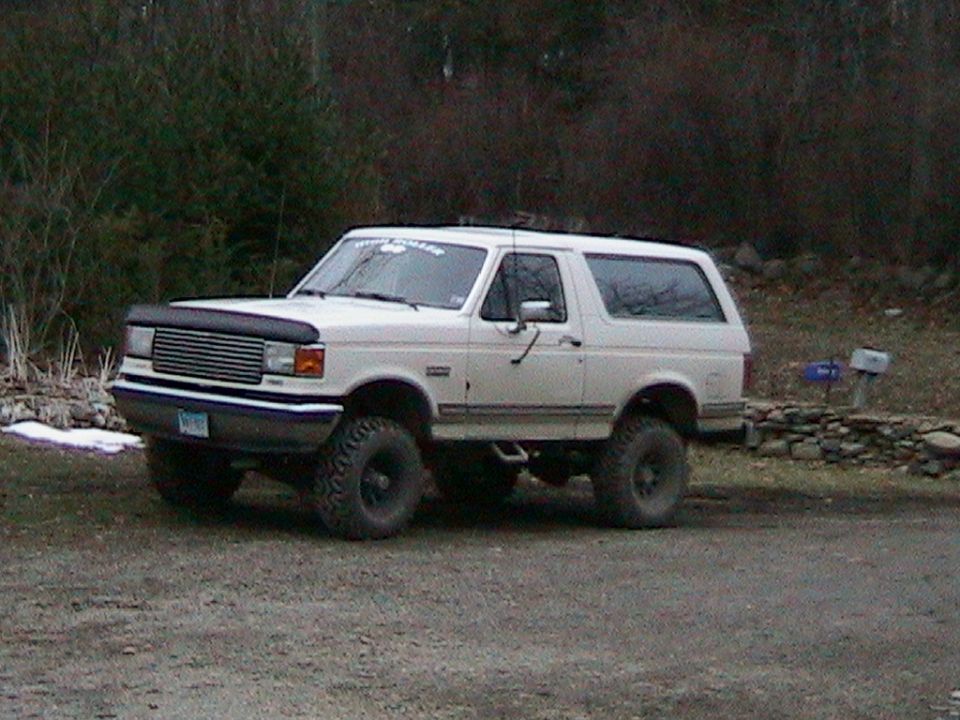 White lifted Ford Bronco with hood bra and roof light bar on gravel driveway