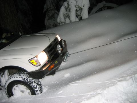 1985 Ford Bronco - Vehicle is stuck in a snowy incline, with only the front visible and headlights illuminated.