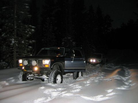 1985 Ford Bronco - Two SUVs navigate a snowy forest at night, one with its door ajar and equipped with off-road tires and a winch.