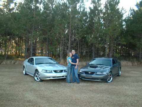 1999 Ford Mustang - Couple poses with their two 1999 Ford Mustangs in front of a wooded area.