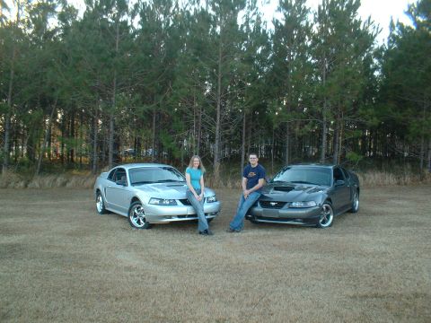 1999 Ford Mustang - Two Ford Mustangs are parked in a grassy field with a line of trees in the background.