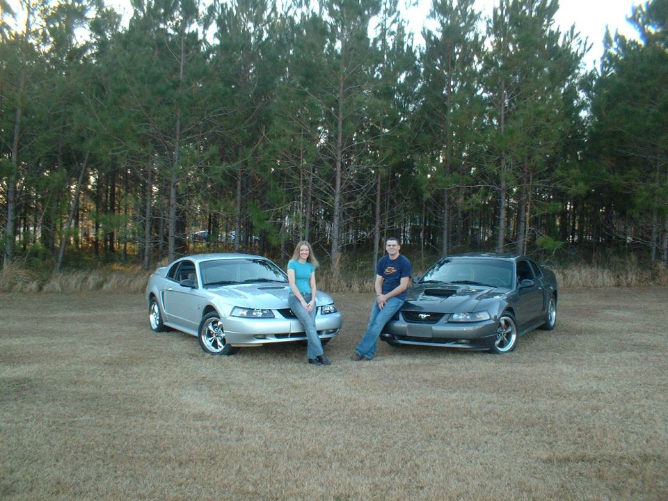 Silver and gray Mustangs parked outside with a man and woman leaning on them
