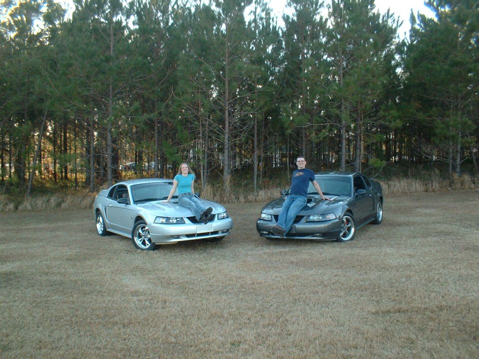 Two 2000 Ford Mustangs, one silver and one gray, parked outdoors with people sitting on hoods