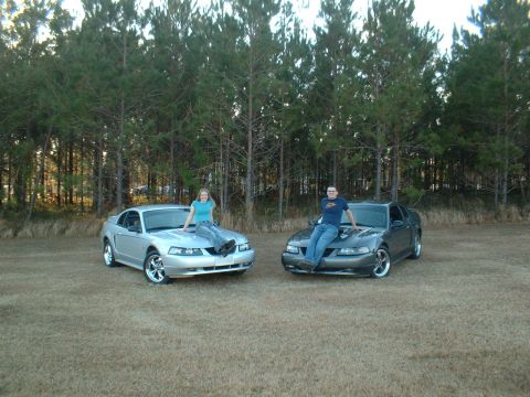1999 Ford Mustang - Two Ford Mustangs are parked side-by-side in a grassy field with trees in the background.
