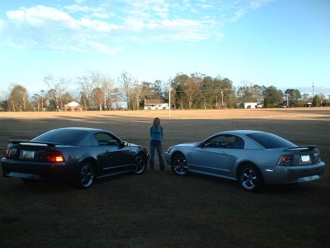 1999 Ford Mustang - Two 1999 Ford Mustangs are parked on a grassy field with a woman standing between them under a partly cloudy sky.