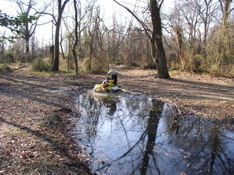 2000 Ford Excursion - An ATV navigates a muddy trail, kicking up water as it goes through a puddle in a wooded area.