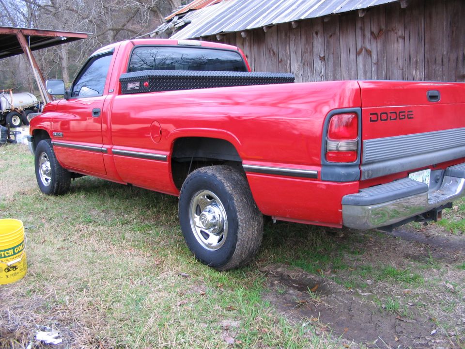 Red Dodge Ram regular-cab pickup with bed toolbox, rear 3/4 view, by a wood barn