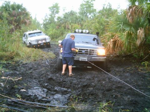 1995 Ford Bronco - Two trucks are stuck in a muddy off-road trail, with one driver attempting to use a winch.