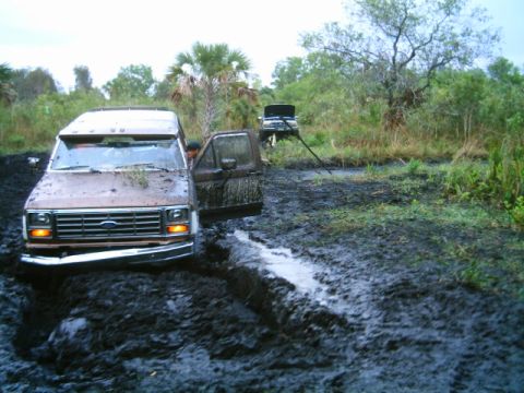 1995 Ford Bronco - Two vehicles are stuck in deep mud with vegetation in the background.
