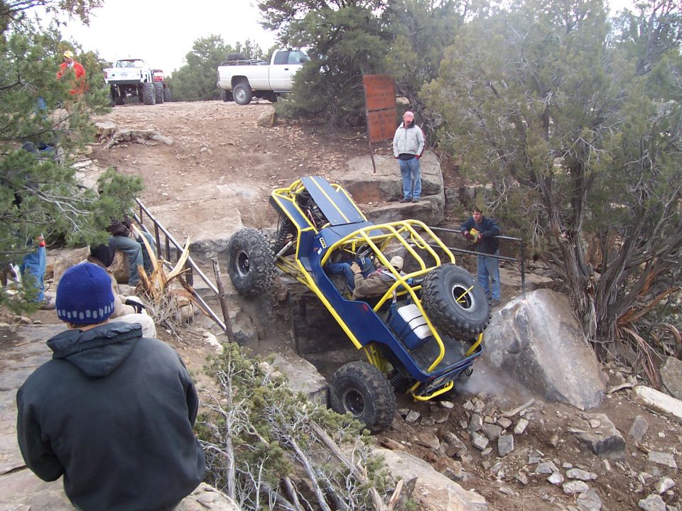 Custom blue and yellow off-road buggy navigating a steep, rocky trail with people watching. White pickup truck visible in background