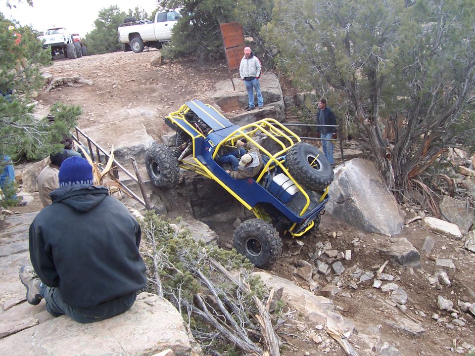 Blue buggy with yellow roll cage climbing rocks. White pickup truck in background