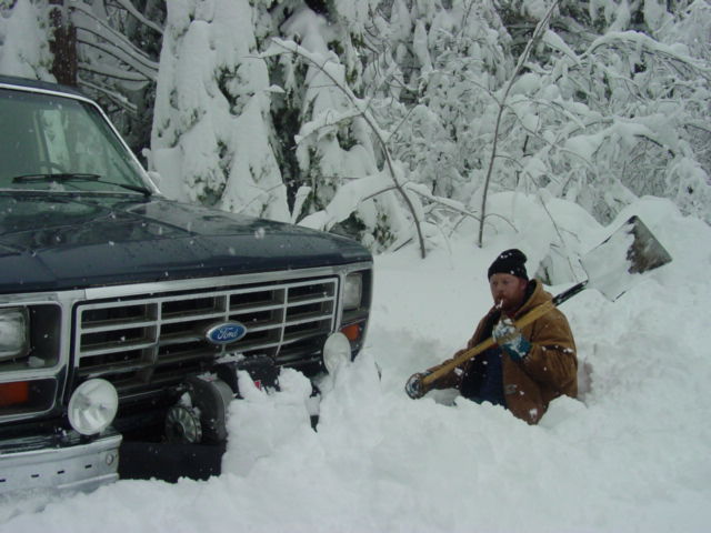 Man in brown coat with shovel sitting in deep snow beside a dark Ford SUV with tire chains during snowfall