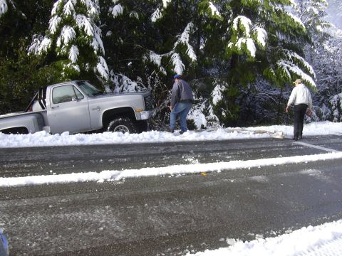 1978 Ford Bronco - A silver pickup truck is stuck in the snow on the side of a road, with two people nearby.