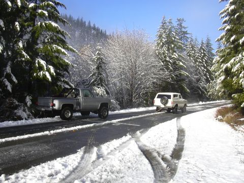 1978 Ford Bronco - Two vehicles navigate a snow-covered road surrounded by pine trees under a clear blue sky.