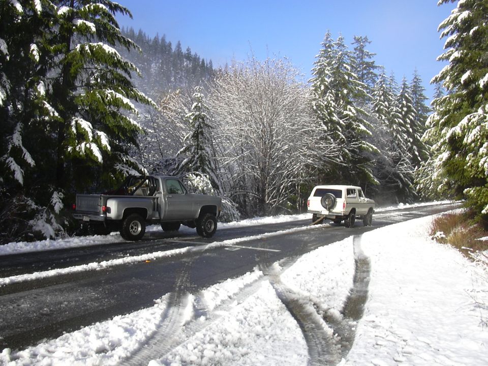 Silver pickup truck and white SUV driving on a snowy road in a forest