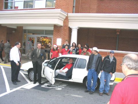 1995 Ford Taurus - A group of people gather around a white sedan parked in front of a building.