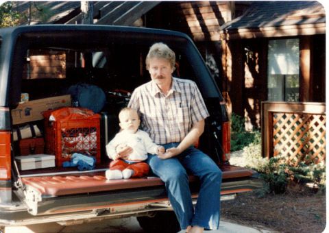 1985 Ford Bronco - Man and baby sit in the open tailgate of a red SUV, surrounded by luggage.