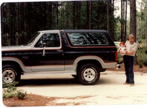 1985 Ford Bronco - Man holds baby next to a 1985 Ford Bronco parked outdoors.