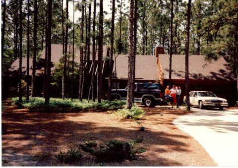1985 Ford Bronco - A dark SUV and a white sedan are parked in the driveway of a house surrounded by pine trees.