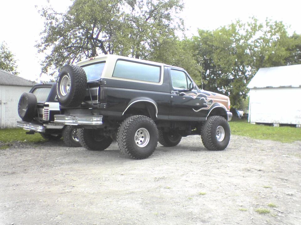 Black and tan lifted Ford Bronco with flame decals and spare tire on rear bumper, parked on gravel