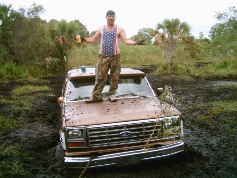 1995 Ford Bronco - Man stands on the hood of a brown SUV stuck in a muddy swamp, holding a beer can.