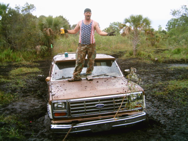 Person standing on the muddy hood of a white Ford truck buried in a swamp mud hole with a tow strap attached