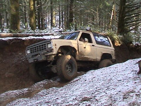 1994 Ford Explorer - A tan Ford Bronco II navigates a challenging muddy and snowy off-road trail through a forest.