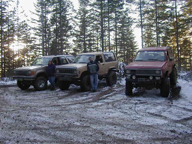 Three lifted 4x4s parked in a snowy pine-forest clearing
